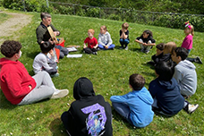 Students sitting in a circle outside on grass listening to a teacher demonstration.