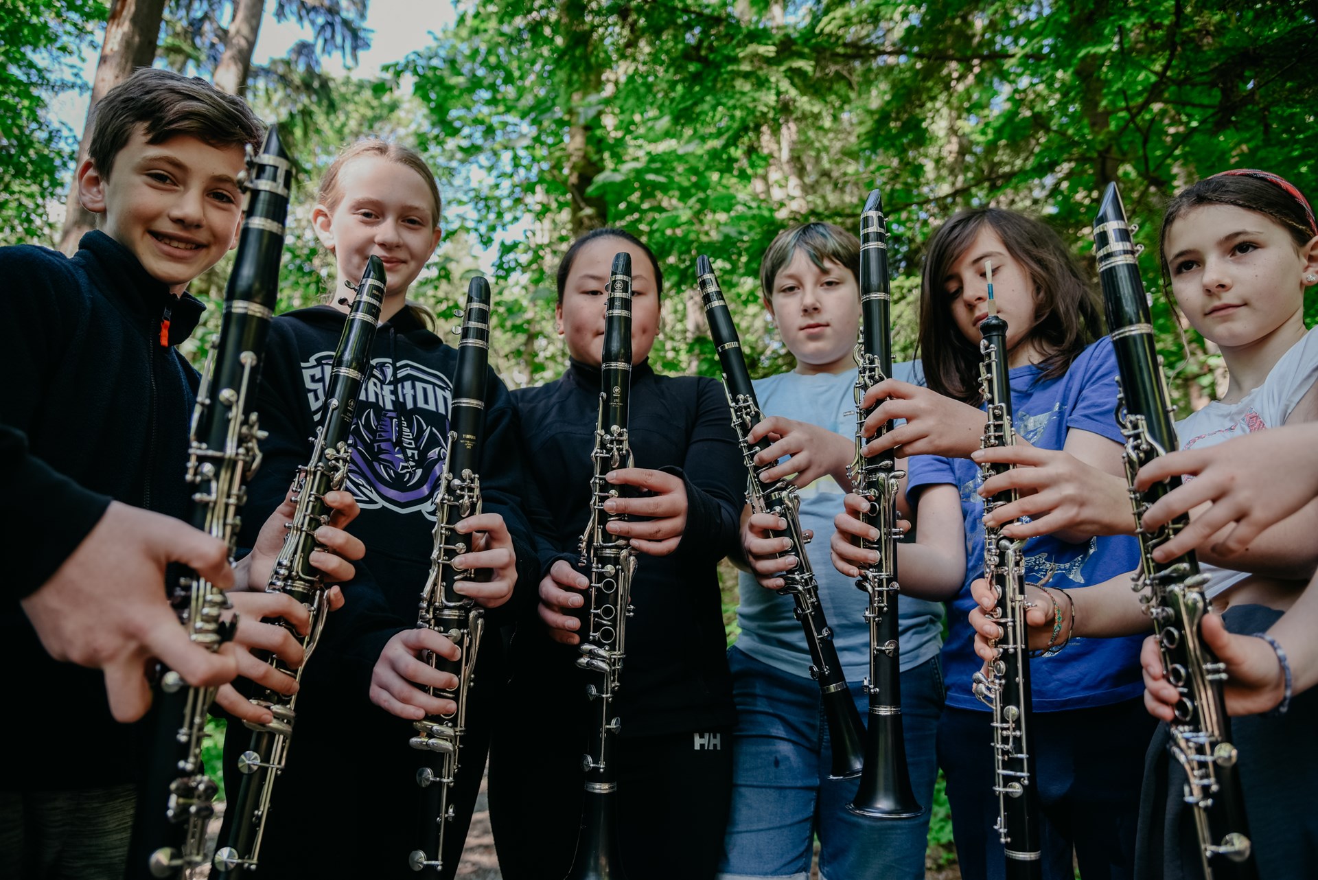Six elementary students playing wind instruments outside in the forest on a sunny day.